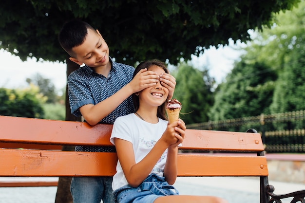 Маленькие друзья на скамейке едят мороженое Мальчик закрыл сестре глаза