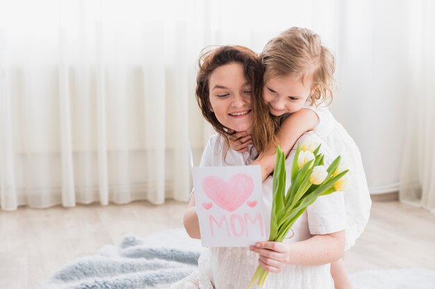 Small girl hugging her mom from back while smiling mother holding greeting card and flowers at home
