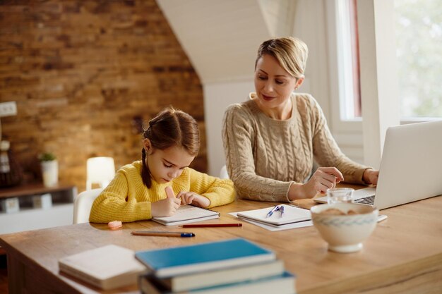 Small girl writing in notebook while her mother is working on laptop at home