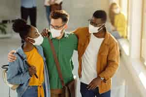 Free photo small group of happy college friends with protective face masks embracing while standing in a lobby and talking.