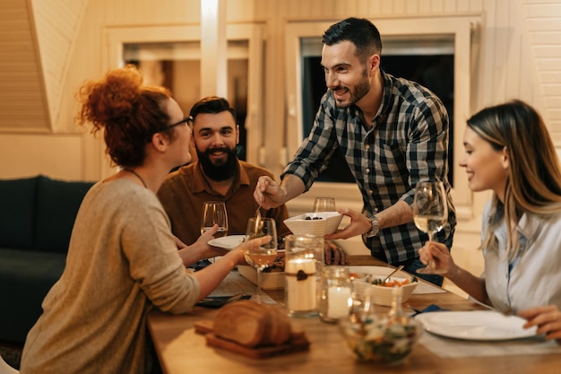 Small group of happy friends eating together at dining table Focus is on man serving food