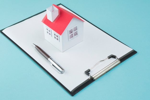 Small house model and pen on blank clipboard over blue backdrop