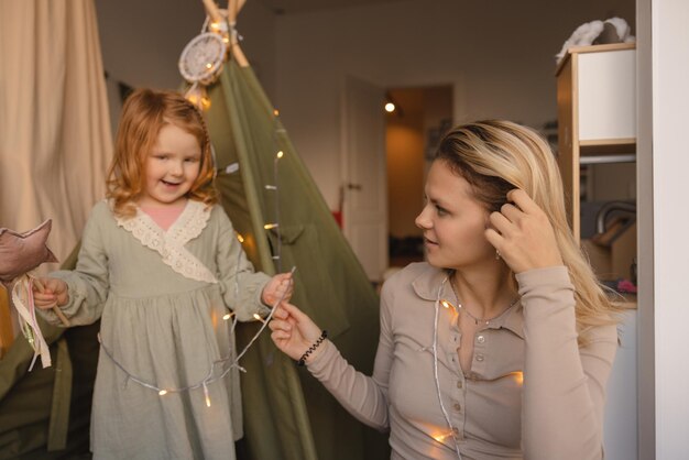 Small lightskinned redhaired girl in dress and young blonde woman are friends while in room Child and mother concept