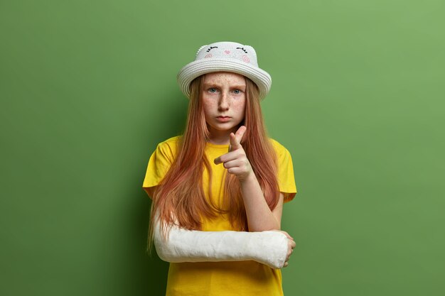 Small little girl with freckled skin and long ginger hair, points at you and looks seriously, wears hat and yellow t shirt, has broken arm after accidental fall, isolated on green wall.