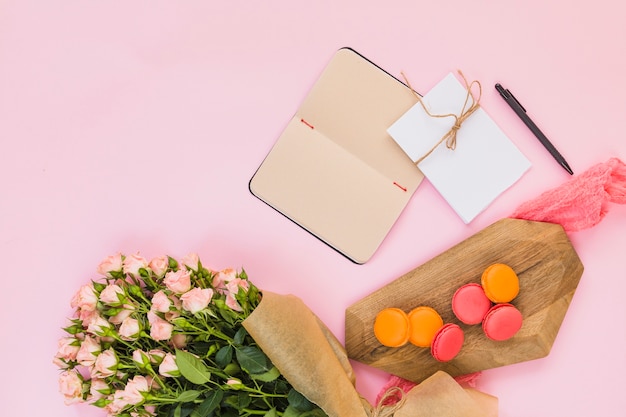 Small macaroons on chopping board; diary; card; pen and flower bouquet against pink background