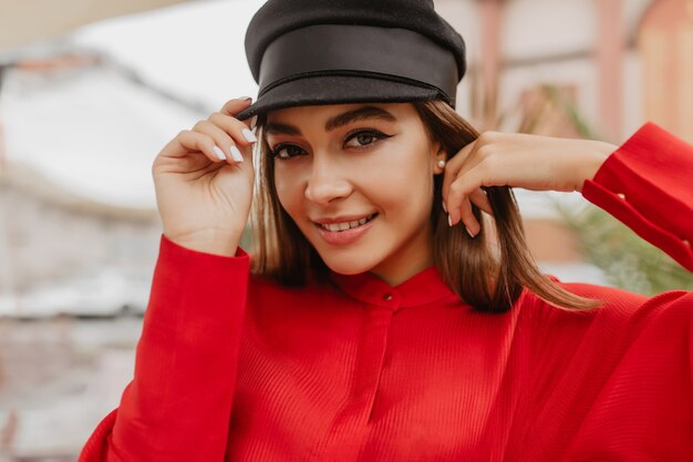 Small pearl earrings attract attention in portrait of young fashion blogger. Girl with sweet smile coquettishly touches her black cap