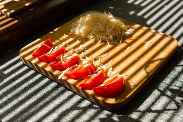 강판에 간 신선한 치즈와 빨간 토마토의 작은 더미는 부엌에 있는 나무 판자에 놓여 있습니다