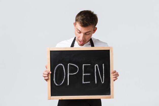 Small retail business owners, cafe and restaurant employees concept. Amused happy salesman, waiter inform about opening, showing open sign and looking upbeat, white background