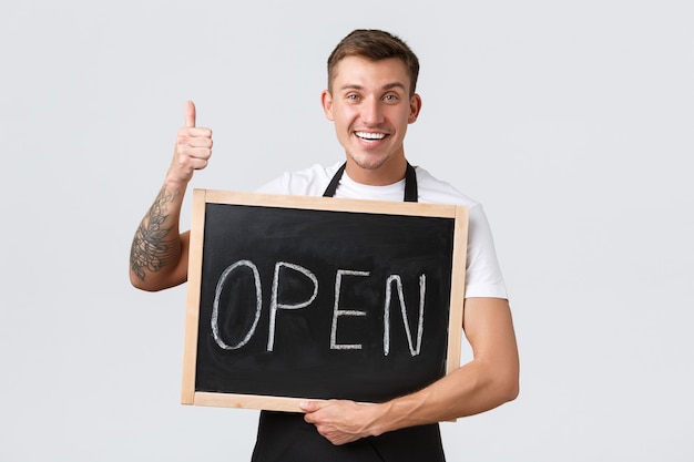 Small retail business owners, cafe and restaurant employees concept. Friendly happy smiling barista, waiter or salesman showing open sign and thumbs-up, inviting to visit store, white background