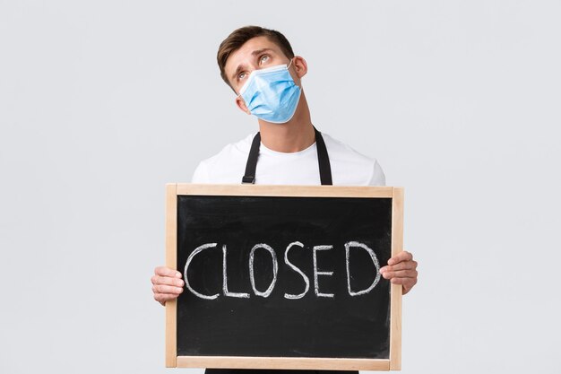 Small retail business owners, covid-19 and social distancing concept. Sad and gloomy salesman, cafe employee in medical mask rolling eyes up and showing closed sign, disappointed with lockdown