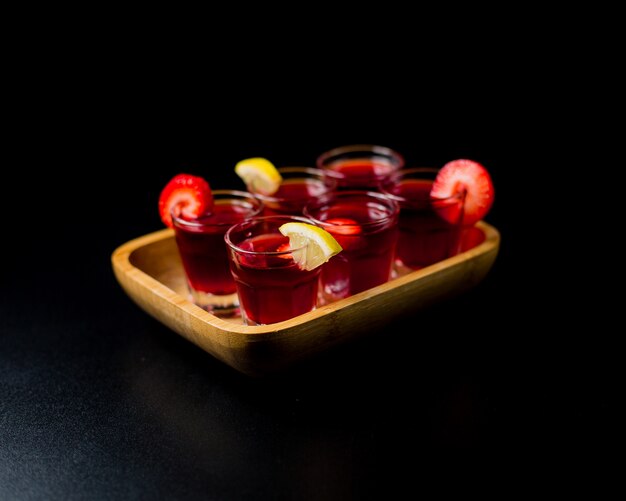 Small sangria glasses with lemon and strawberries inside bamboo platter in a black space.