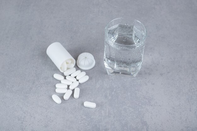 A small white jar of white pills with glass of pure water on gray surface