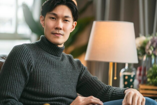Умная привлекательная азиатская мужская стильная дизайнерская улыбка с уверенной и веселой домашней мужской свитер сидит на диване в гостиной дома