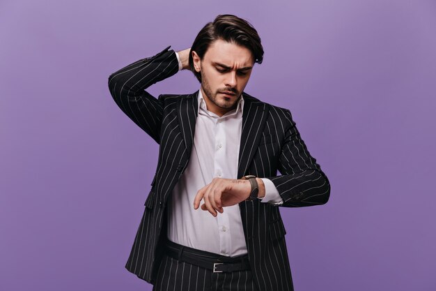 Smart attractive man with brunette hair, white shirt and trendy striped suit, looking at watch on his hand