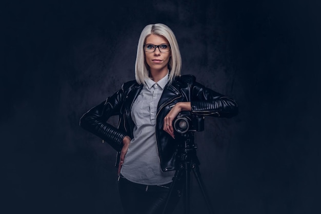 Smart blonde female photographer in trendy clothes posing while leaning on a professional camera with a tripod at a studio, looks at the camera. Isolated on a dark background.