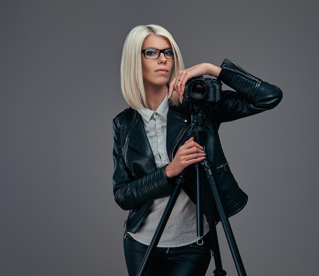 Fotografa bionda intelligente in abiti alla moda in posa mentre si appoggia su una fotocamera professionale con un treppiede in uno studio, guarda la fotocamera. isolato su sfondo grigio.