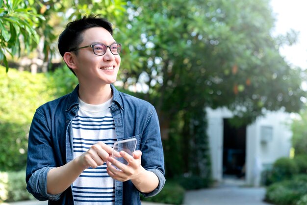 똑똑하고 쾌활한 아시아 안경 남성 손은 아름다운 녹색 나무 정원 홈 개념의 배경과 투명 유리 스마트폰 미래 기술 연결을 사용합니다