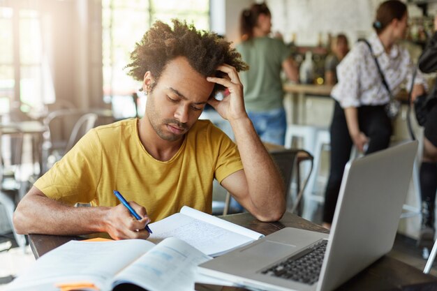 Smart college student in casual clothes looking attentively in his notebook writing notes using laptop computer being focused at his writing sitting in coffee shop. Hard-working male being busy