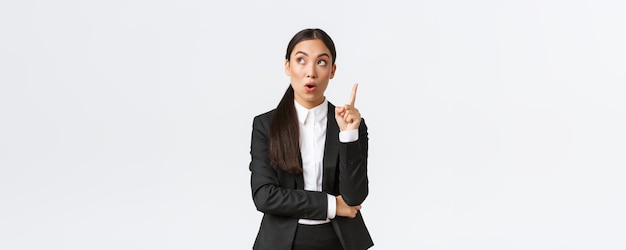 Smart and creative asian businesswoman have excellent idea raising finger up and looking upper left corner thoughtful suggest solution make plan saying thought over white background