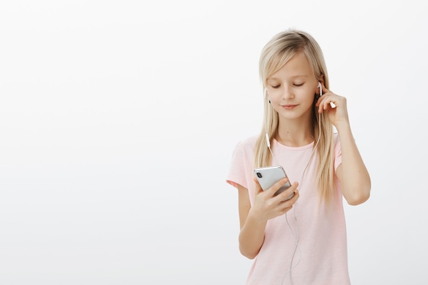 Smart girl knows everything about gadgets. Portrait of cute beautiful blond young child, wearing earphones and picking song in smartphone