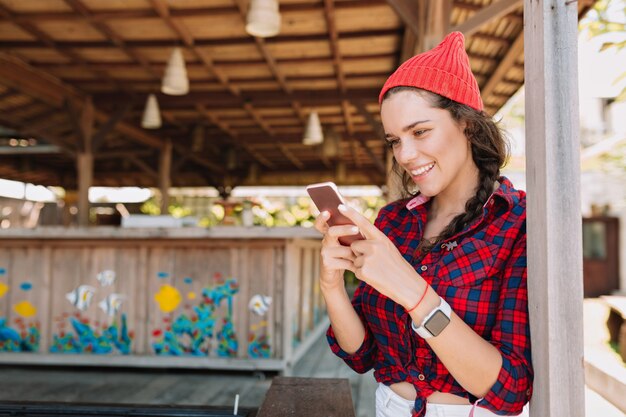 日光の下で愛らしい笑顔でスマートフォンでスクロールする素敵な笑顔を持つスマートな流行に敏感な女性。外のスマートフォンで白い歯と健康な肌を持つ女性