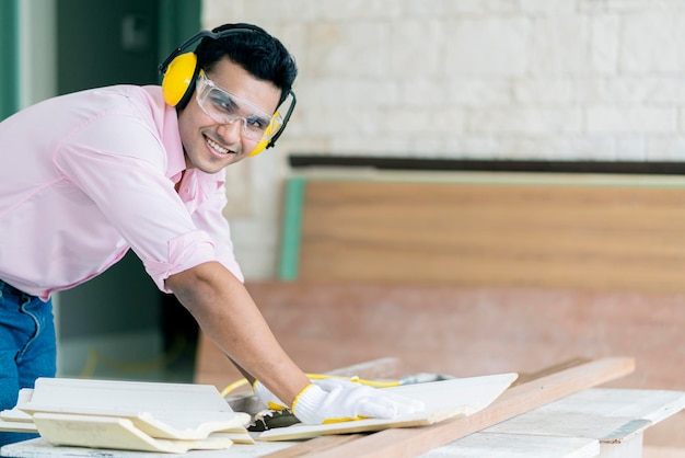 Smart indian carpenter contractor working with sawing machine with piece of moulding cornice and plank wooden part of built in furniture