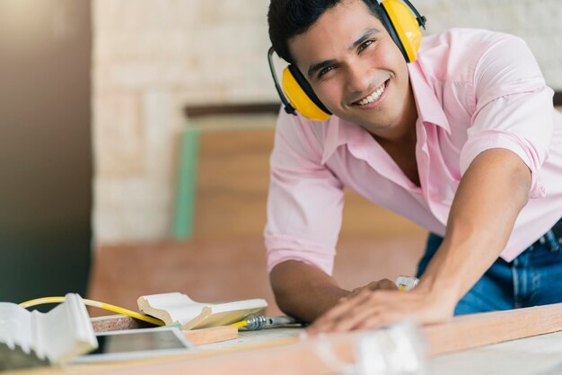 Smart indian carpenter contractor working with sawing machine with piece of moulding cornice and plank wooden part of built in furniture