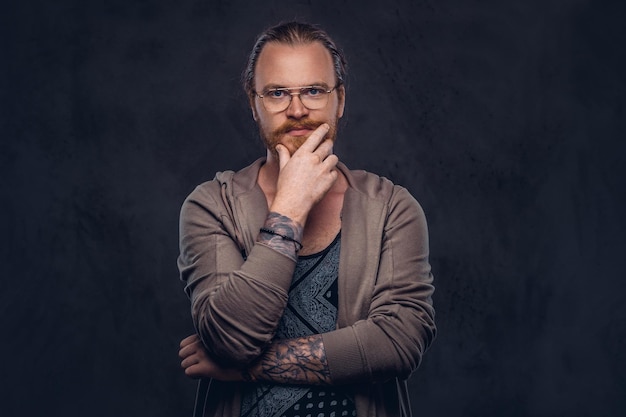 Free photo smart pensive redhead hipster with full beard and glasses dressed in casual clothes, poses with hand on chin in a studio. isolated on the dark background.