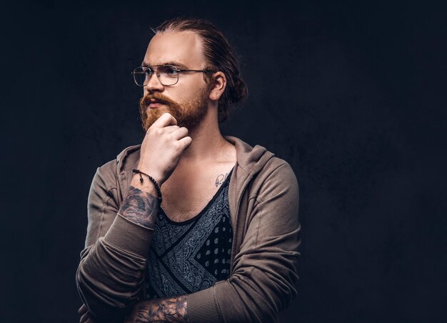 Smart pensive redhead hipster with full beard and glasses dressed in casual clothes, poses with hand on chin in a studio. Isolated on the dark background.