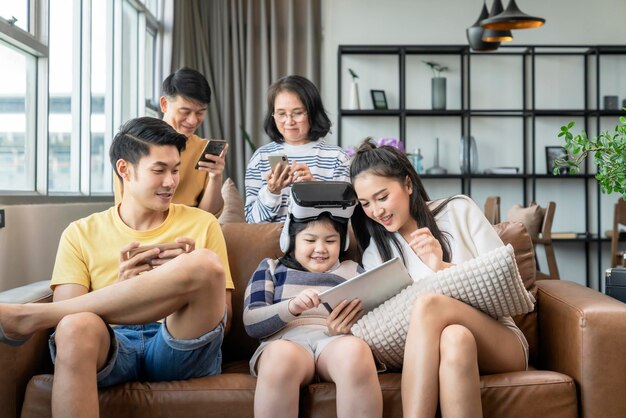 ホームアジア世代の家族のライフスタイルとスマートテクノロジーは、自宅の検疫の瞬間にソファのリビングルームで一緒に楽しく遊ぶスマートフォンタブレットとvrゴーグルメガネを使用してテクノロジーデバイスをお楽しみください