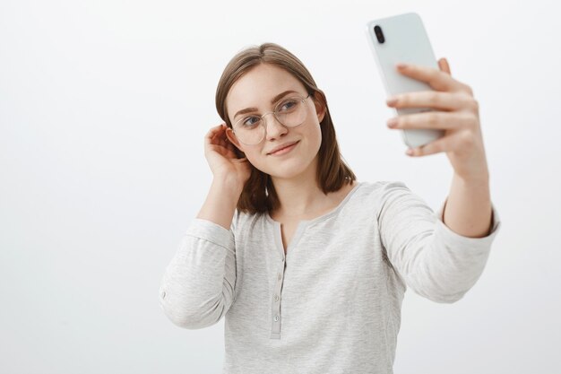 Умная и нежная женщина в очках, откидывающая прядь волос за ухом и мило улыбаясь, делая селфи на новеньком смартфоне, позирует над серой стеной, довольная, делая пост в социальной сети