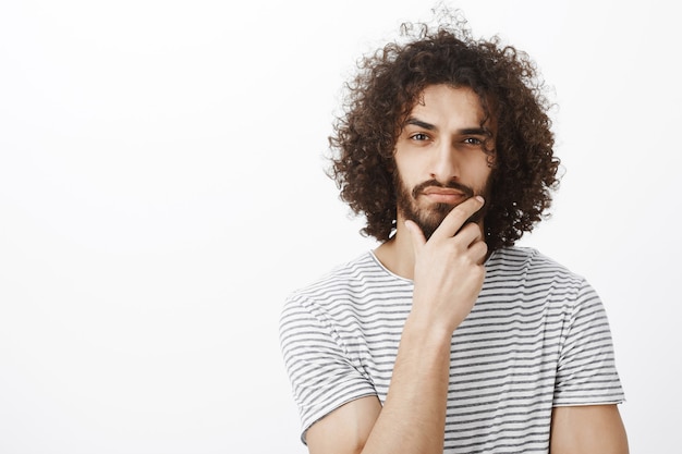 Smart thoughtful attractive hispanic guy with beard and curly hair, holding hand on chin, thinking