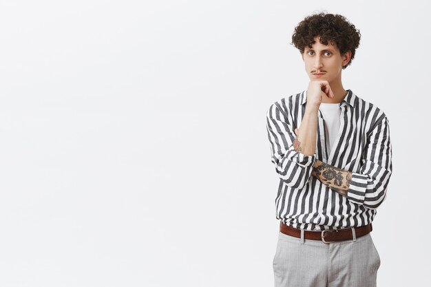 Smart and thoughtful young creative male entrepreneur with tattooed arm moustache and curly hair in trendy formal striped shirt and pants leaning on fist and looking seriously