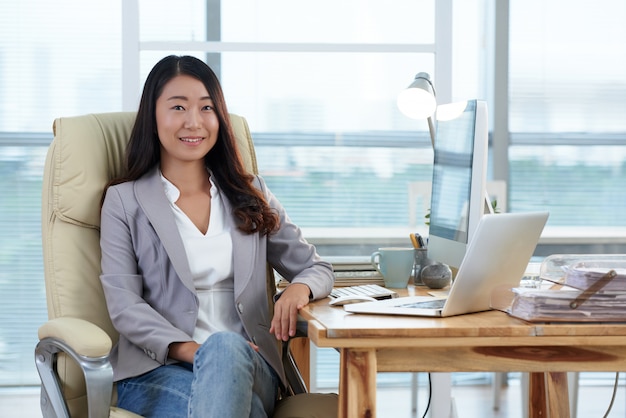 Smartly dressed Asian lady sitting in office with computer and laptop and smiling