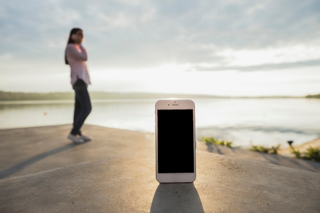 Smartphone in front of woman talking on cellphone standing near the lake