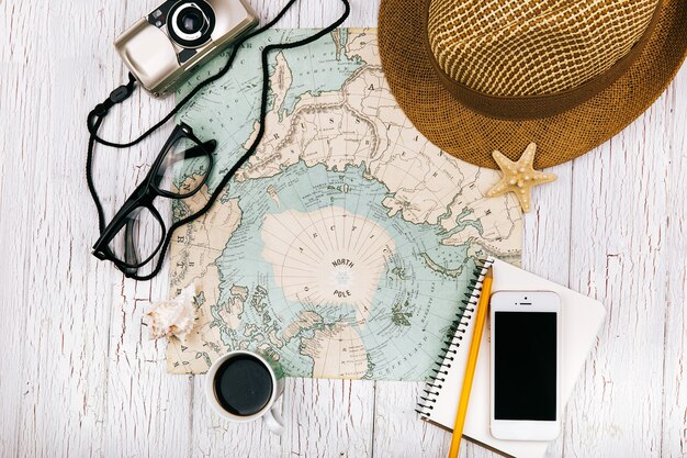 Smartphone lies on a notebook before a cup of coffee on the map, hat, camera and glasses around it