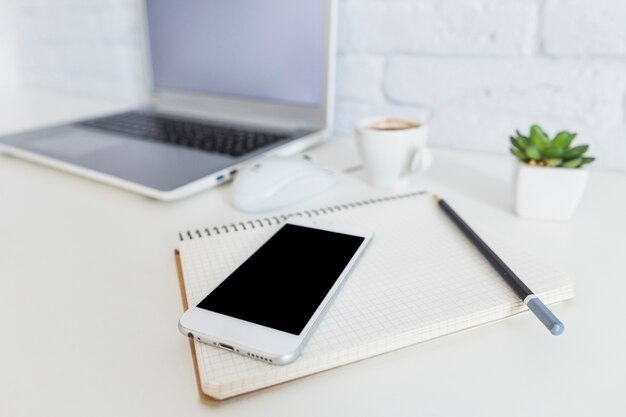 白い机の上のラップトップの前にスマートフォン、ノート、鉛筆