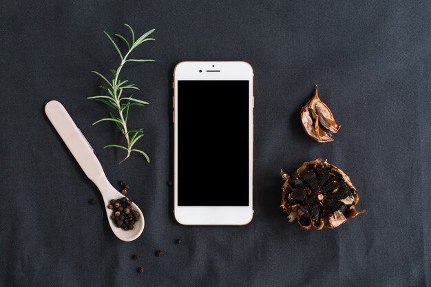 Smartphone surrounded with rosemary; black pepper and garlic on dark backdrop