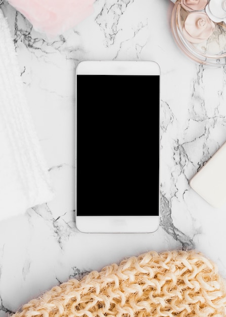 Smartphone surrounded with scrub glove; soap; perfume bottle; towel and sponge on marble backdrop