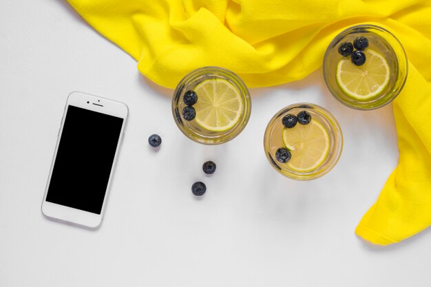 Smartphone with lemon juice glasses and yellow textile on white backdrop