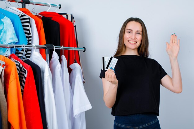 Smiing girl is holding credit cards and showing hi gesture with other hand on clothes background