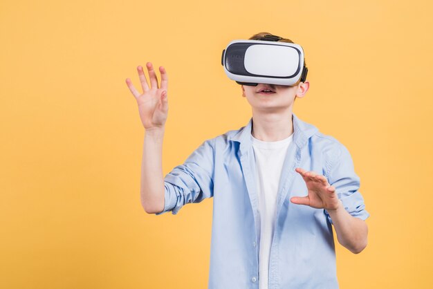 Smile boy wearing using virtual reality glasses headset moving his hands in air against yellow background