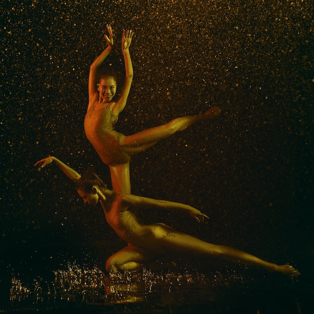 Smile. Two young female ballet dancers under water drops and spray. Caucasian and asian models dancing together in neon lights. Ballet and contemporary choreography concept. Creative art photo.