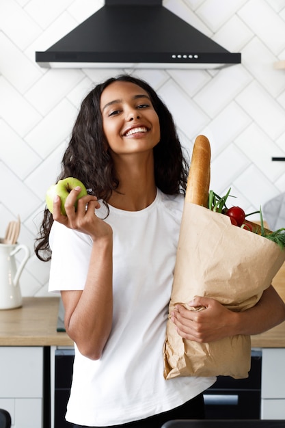 Free photo smiled beautiful mulatto woman is holding package full with food in one hand and apple in other on the modern white kitchen