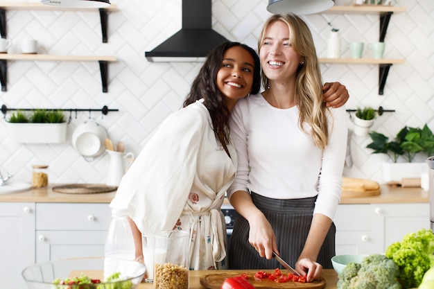 Smiled blonde caucasian woman is cutting pepper and brunette mulatto woman is hugging her on the white modern kitchen