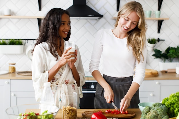 Smiled blonde caucasian woman is cutting pepper and brunette mulatto woman in nightwear is drinking milk and looking on her on the white modern kitchen