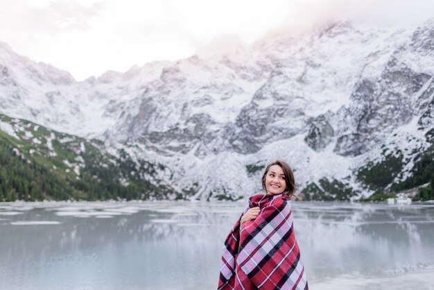 暖かい毛布で覆われている微笑んでいる女の子は山、若い旅行者の湖の近くに立っています。
