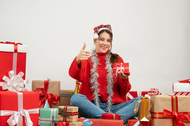 smiled girl with santa hat holding present and making thumb up sign sitting around presents on white