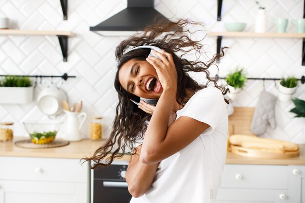 Smiled hilarious mulatto woman with messy hair in big wireless headphones is happily dancing with her opened mouth in the modern kitchen