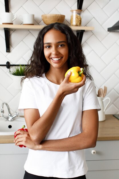 Улыбчивая женщина-мулатка, одетая в белую футболку, с красивым лицом и распущенными волосами, держит в руке желтый перец возле кухонного стола на современной кухне